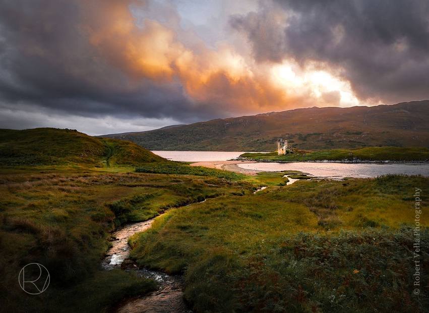 A loch with a ruined castle on the shore, sunlight pushing through the clouds behind it